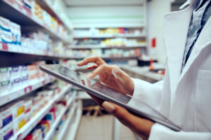 Pharmacy technician taps an iPad standing in front of a shelf of medications.